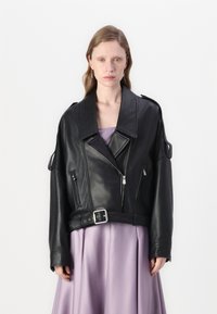 Jaqueta cropped de couro preto com lapelas largas, bolsos com zíper e cintura com cinto. Usada sobre um vestido lilás claro com um design com pregas.
