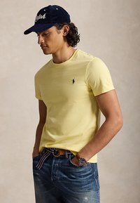 Mann som har på seg en gul t-skjorte, blå jeans, en marineblå cap og armbåndsur, med hendene i lommene, ser ned til siden.
