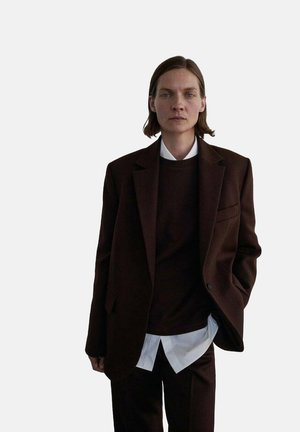 Blazer marrone sartoriale indossato su un maglione scuro, abbinato a una camicia bianca con bottoni e pantaloni coordinati. Tessuto liscio, design strutturato.