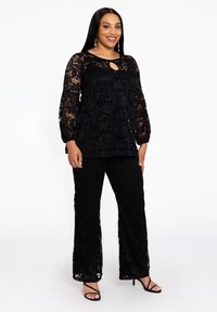 Zwarte kanttop met lange, doorschijnende mouwen en een sleutelgatdetail, gecombineerd met bijpassende zwarte kantbroeken, beide met een bloemenpatroon.