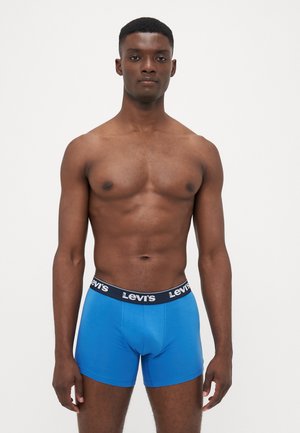 Man som står vänd framåt, klädd i blå Levi's kalsonger med svart midjeband mot en enkel vit bakgrund.