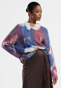 Maglione multicolore lavorato a maglia, con tonalità di blu, rosa e crema, dal taglio rilassato e colletto rotondo, abbinato a una gonna marrone con frange.