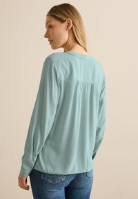 Blouse bleu clair à manches longues avec une coupe décontractée, tissu texturé, et des détails froncés à l'arrière. Associée à un jean bleu foncé.