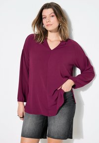 Burgunderfarbene Bluse mit langen Ärmeln und V-Ausschnitt, strukturierter Stoff und gerafften Ärmeln, kombiniert mit dunklen Denim-Shorts. Schlichtes, lässiges Design.