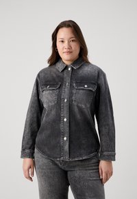 Črna denim srajca z dvema prsne žepoma, srebrnimi gumbi na zapenjanje in rahlo zbledelim izgledom, v kombinaciji s temnimi kavbojkami.