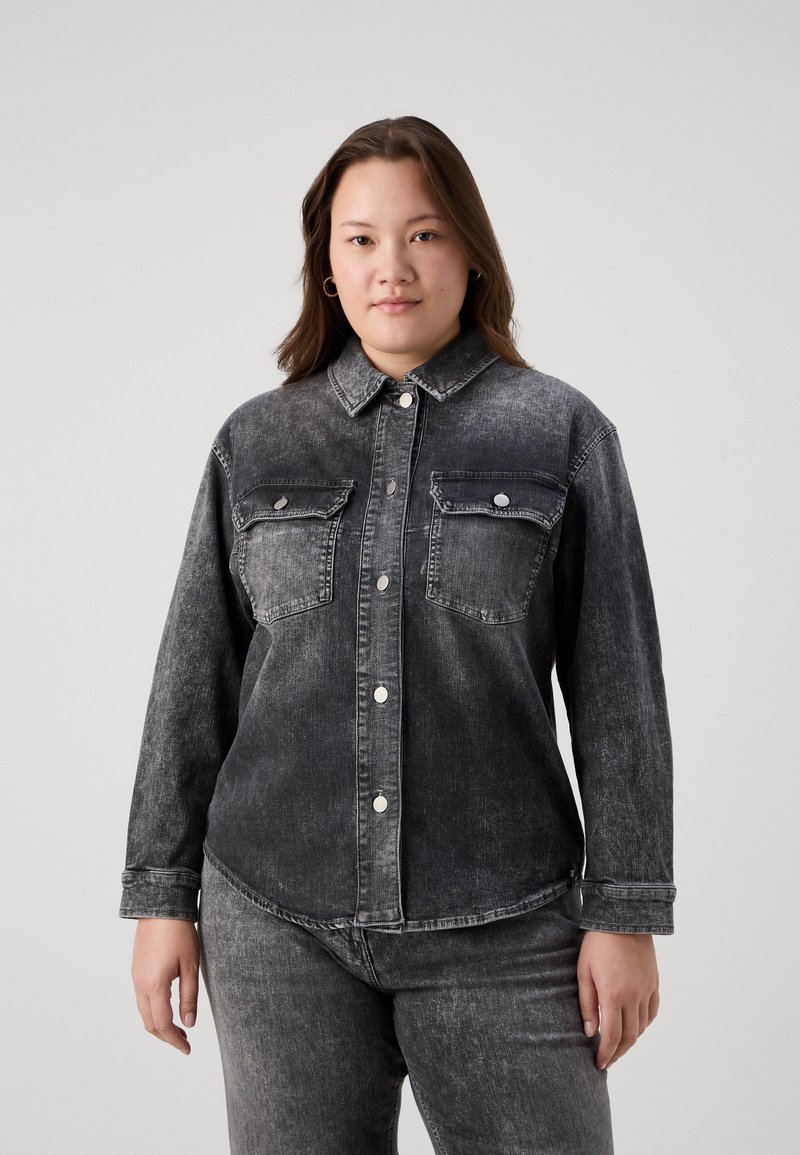 Črna denim srajca z dvema prsne žepoma, srebrnimi gumbi na zapenjanje in rahlo zbledelim izgledom, v kombinaciji s temnimi kavbojkami.