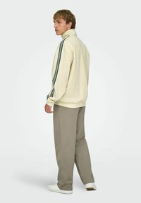 Jeune homme debout, portant une veste zippée crème avec des rayures noires sur les manches, un pantalon taupe et des baskets blanches, faisant face à gauche.