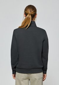 Svart sweatshirt med hög krage, ribbade ärmmuddar och en slät yta, tillsammans med ljusa byxor.