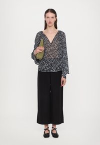Blouse noire à fleurs avec manches longues, associée à un pantalon large noir, tenant un sac à main vert ; tissu texturé, coupe décontractée.
