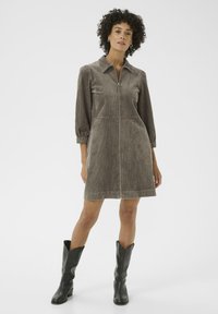 Abito in corduroy marrone con colletto, cerniere e maniche a tre quarti. Presenta un corpino aderente e una gonna svasata, abbinato a stivali neri.