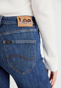 Jeans in denim blu scuro con patch in pelle con logo Lee, cuciture gialle e design delle tasche posteriori curvo. Il materiale sembra texturizzato e resistente.