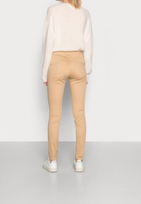Beige jeans med åtsittande passform och bakfickor, i kombination med en ljus, texturerad tröja. Modellen står på ett grått golv och har på sig vita sneakers.