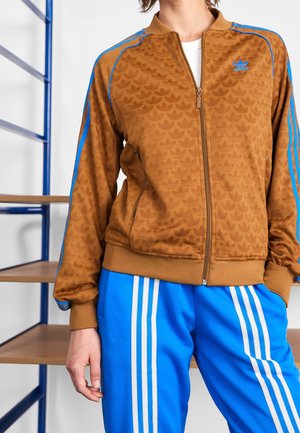 Persoon die een bruine Adidas-jas met blauwe strepen en blauwe broek met witte verticale strepen draagt, staand naast houten planken.