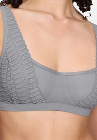 Bralette grigio con rifiniture in pizzo strutturato, tessuto liscio e spalline larghe e piatte. Il design presenta un scollo poco profondo e non ha ferretto.