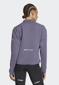Chaqueta texturizada de color púrpura con acentos reflectantes, mangas largas y un corte relajado. Combinada con leggings negros, mostrando un diseño deportivo.