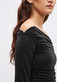 Zwarte top met lange mouwen en off-shoulder ontwerp, met geplooide textuur aan de voorkant en gladde stof. Subtiele glans in het materiaal.