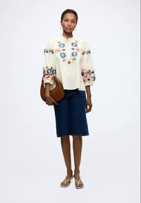 Femme portant un chemisier crème avec une broderie florale colorée, un short en jean jusqu'aux genoux, des sandales blanches, tenant un sac à main marron.