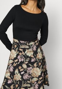 Svart långärmad topp i kombination med en blommig kjol som har stora blommor i rosa och kräm mot en svart bakgrund. Släta texturer, figurnära siluett.