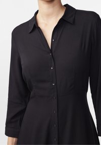 Donna che indossa un vestito nero con bottoni, colletto e maniche a tre quarti, mostrata dalla gola alla vita su uno sfondo bianco.
