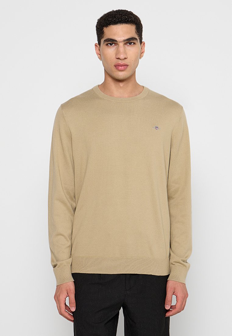 Beige Strickpullover mit Rundhalsausschnitt, langen Ärmeln und kleinem gesticktem Logo auf der Brust. Glatte Textur und figurbetontes Design.