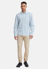 Helles blaues Hemd mit Knopfleiste und Brusttasche, kombiniert mit beigen Hosen und schwarzen Schuhen, vor einem schlichten weißen Hintergrund präsentiert.