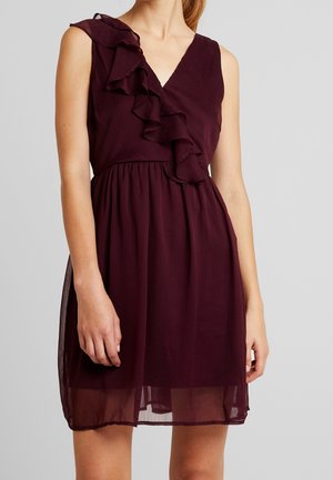 Burgunderfärgad ärmlös klänning med V-ringning och volangdetalj längs ena axeln. Tyget är lätt och har en svävande kjol.