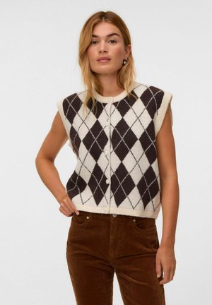 Vrouw draagt een mouwloos zwart-wit argyle gebreid vest en een bruine corduroybloes, staat met hand op heup.