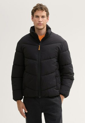 Chaqueta acolchada negra con cuello alto, cierre de cremallera y acentos en naranja. Presenta patrones de costura geométrica y puños elásticos. Corte casual.