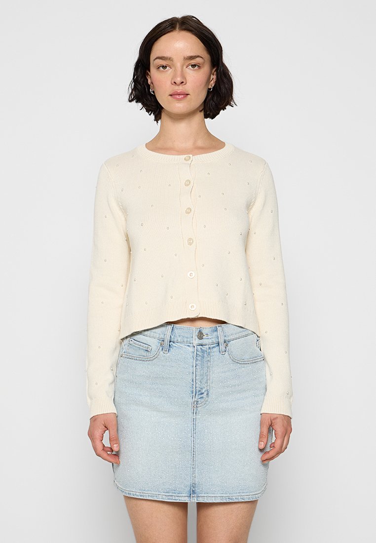 Cremefarbener, taillierter Cardigan mit Knopfverschluss, strukturiert mit kleinen, erhabenen Punkten, kombiniert mit einem hellen Jeansrock.