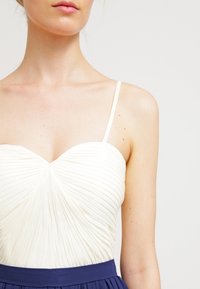 Biała pleciona bluzka z dekoltem w kształcie serca i cienkimi ramiączkami, w połączeniu z granatową spódnicą. Materiał wydaje się gładki i lekki.