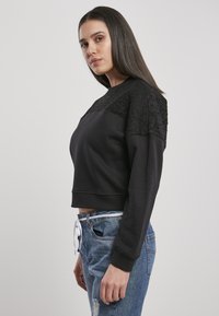 Felpa nera corta con dettagli in pizzo testurizzato sulle spalle, polsini a coste e scollatura rotonda, abbinata a jeans blu strappati.