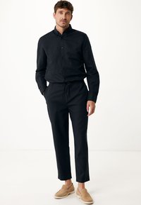 Svart långärmad skjorta med knappar och krage, ihop med matchande byxor med dragsko. Modellen bär beige slip-on skor.