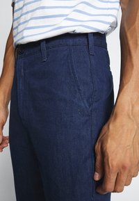 Tummansinisiä farkkuhousuja, joissa on suora lahje, sivutaskut ja napitus vyötäröllä. Kestävä ja teksturoitu kangas.