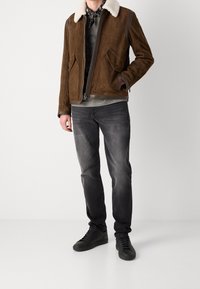 Homme portant une veste en daim marron avec un col en peau lainée blanche, un jean gris délavé, des baskets noires et un bandana noir autour du cou.