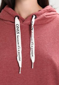 Lähikuva henkilöstä, joka pitää yllä punaista hupparia, jossa on valkoiset nyörit ja teksti "VAIN koska se on intohimo."