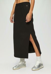 Zwarte midi-rok van zachte katoen, met een hoge taille met elastiek, zijsplitten en een subtiele logo-details. Gecombineerd met zilveren sneakers.