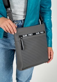 Persona con giacca blu e jeans che tiene una borsa a tracolla grigia a costine con cerniera nera e il testo "SURI FREY" sulla tracolla.