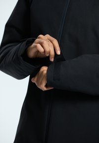 Svart vattentät jacka med mjuk textur, utrustad med dragkedja och justerbara manschetter som säkras med kardborre. Fokus på hand som justerar manschetten.