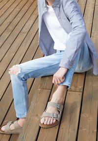 Light grey sandals with two adjustable straps and metal buckles, worn with light blue distressed jeans on a wooden surface.