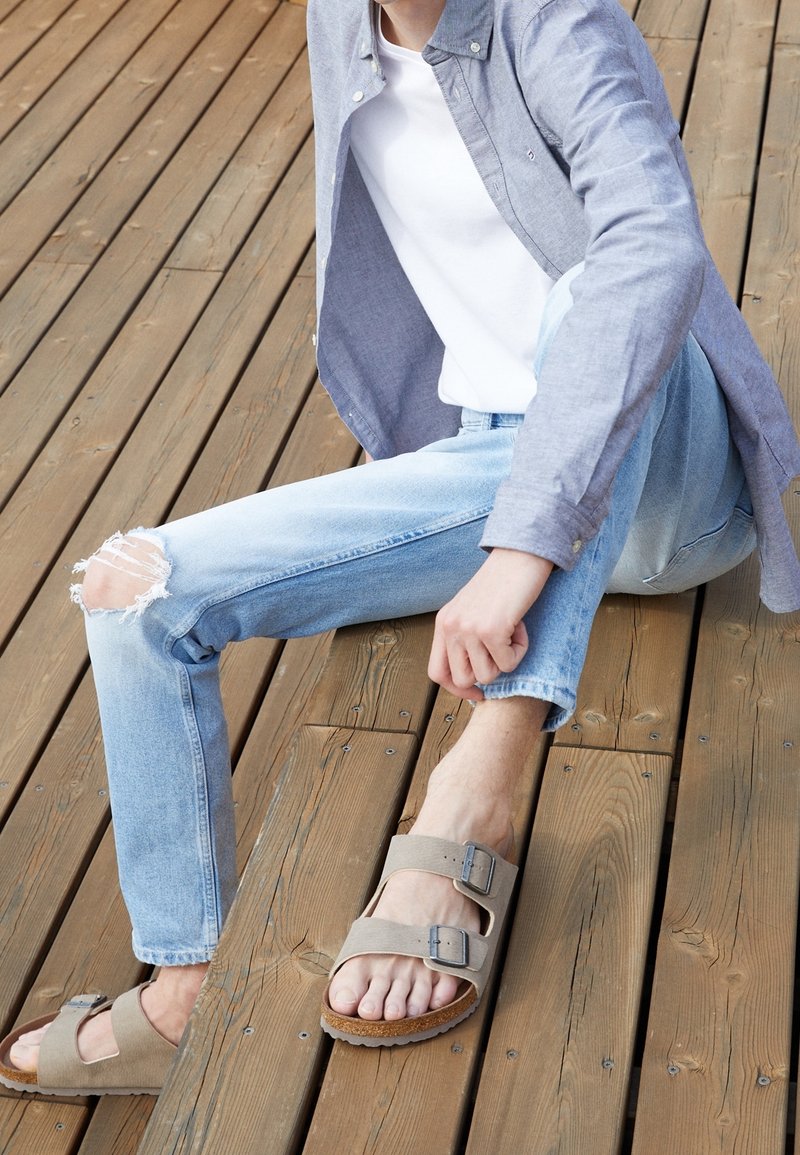 Light grey sandals with two adjustable straps and metal buckles, worn with light blue distressed jeans on a wooden surface.