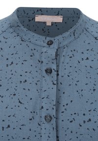 Camicia azzurro chiaro con un motivo testurizzato caratterizzato da macchie nere sparse. Colletto rotondo, tre bottoni neri e un tessuto morbido.