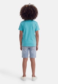 Παιδί με σγουρά μαλλιά στέκεται με την πλάτη γυρισμένη, φοράει γαλάζιο μπλουζάκι, ανοιχτόχρωμα τζιν σορτς και λευκά παπούτσια χωρίς κορδόνια.