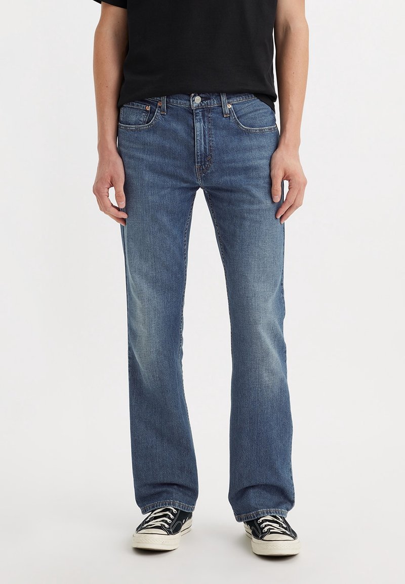 Jeans de mezclilla azul con corte recto, con un ligero desgaste, diseño de cinco bolsillos y herrajes metálicos. Combinados con zapatillas negras.