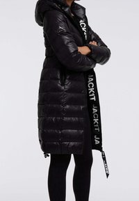 Persona che indossa un lungo piumino nero, lucido e trapuntato, con cappuccio, braccia incrociate e visibile branding bianco "JACK1T" sulla cerniera e sulla manica.