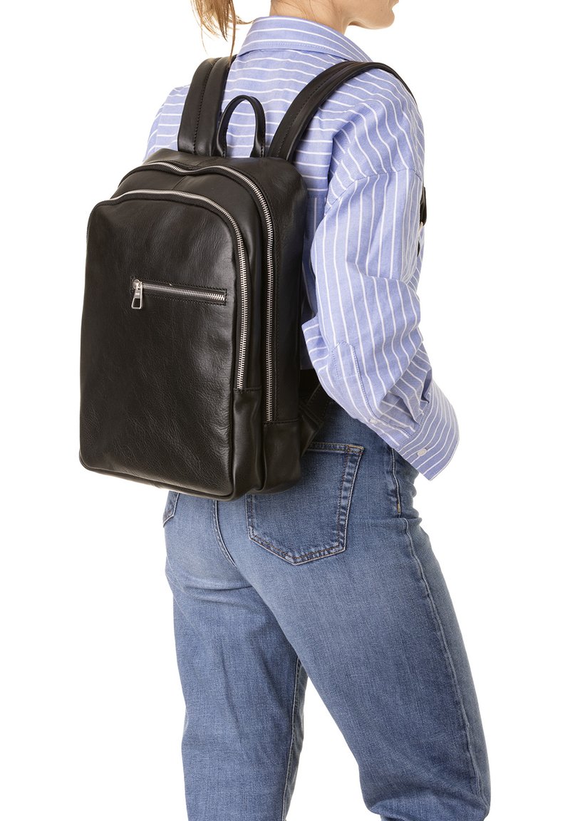 Sac à dos en cuir noir avec deux compartiments zippés, des bretelles ajustables et une texture lisse, porté sur le dos d'une personne en tenue décontractée.