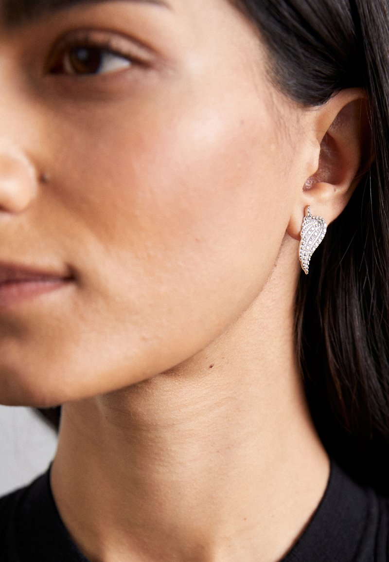 Sudraba lapu formas auskari ar teksturētu dizainu, kuros ir mazi caurspīdīgi dārgakmeņi, kas valkāti modelei uz auss pret neitrālu fonu.