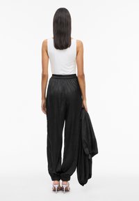 Czarne satynowe spodnie z elastycznym pasem i mankietami na końcach nóg, zestawione z dopasowanym białym topem na ramiączkach. Czarna kurtka zarzucona na ramię.