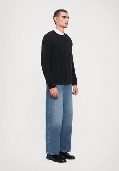 Black cable-knit sweater, blue wide-leg jeans, and black shoes. The sweater has a textured pattern; jeans are high-waisted with a relaxed fit.
