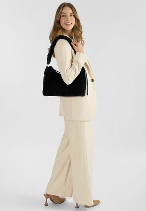 Borsa nera in pelliccia sintetica con tracolla imbottita, forma rettangolare e chiusura con cerniera. Indossata con un completo beige chiaro e tacchi.