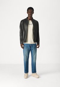 Giubbotto in pelle nera con finitura liscia, t-shirt beige con stampa, jeans di denim blu e scarpe di colore chiaro. Il modello è rivolto in avanti.